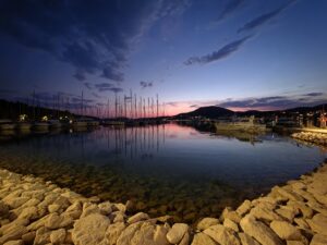 Yacht harbour at sunset with rocks on the foreground and boats with masts in the background. Sunset giving off a pink hue to a dark blue sky