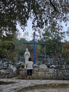 A small outdoor shrine on a rocky hillside, featuring a blue cross and a statue of the Virgin Mary surrounded by flowers and prayer offerings.