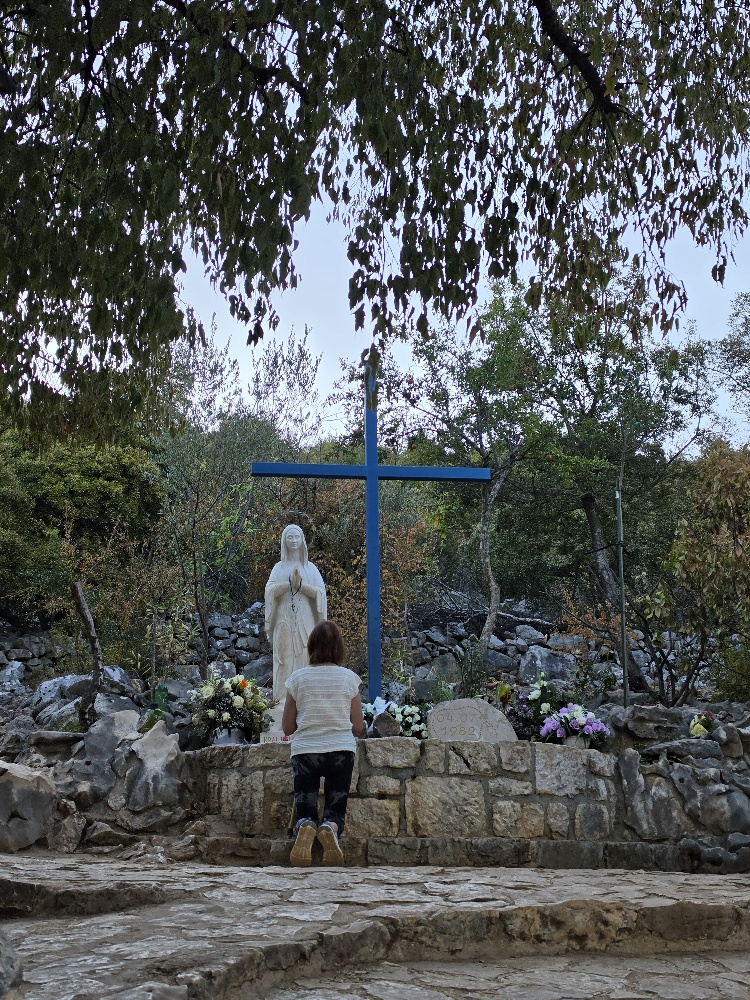A small outdoor shrine on a rocky hillside, featuring a blue cross and a statue of the Virgin Mary surrounded by flowers and prayer offerings.