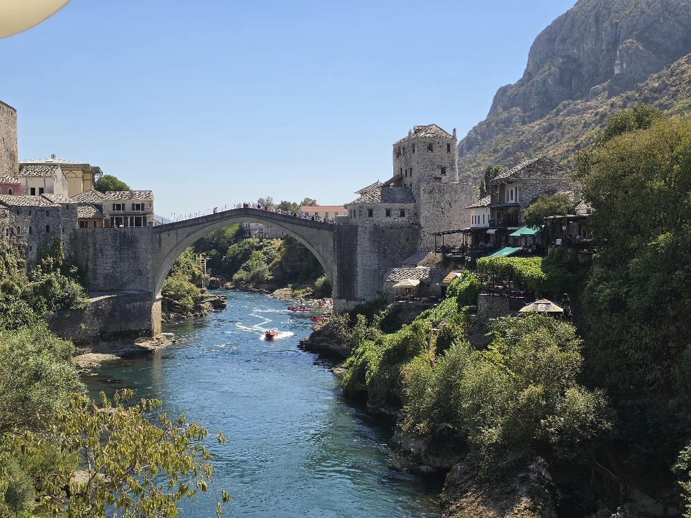 View of Mostar's reconstructed Old Bridge spanning the Neretva River, with historic stone buildings and a tower on either side.