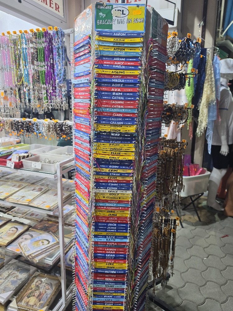 A religious souvenir shop displaying colorful name bracelets, rosaries, prayer cards, religious paintings, and other devotional items