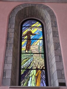 Stained-glass window depicting St Francis of Assisi standing with outstretched arms beneath rays of light, with a crowd of people gathered below.