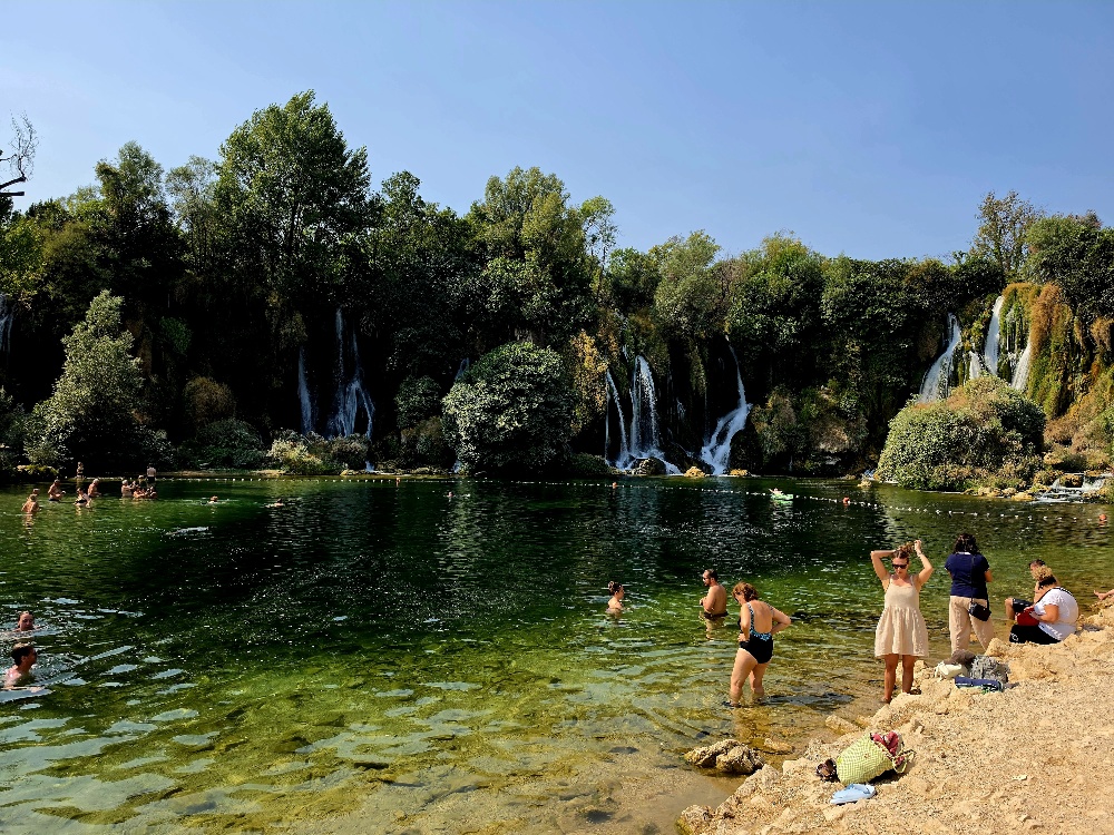 “Kravica Falls on the Trebižat River, with multiple cascades surrounded by lush trees and moss-covered limestone, and visitors swimming in the pool below.”