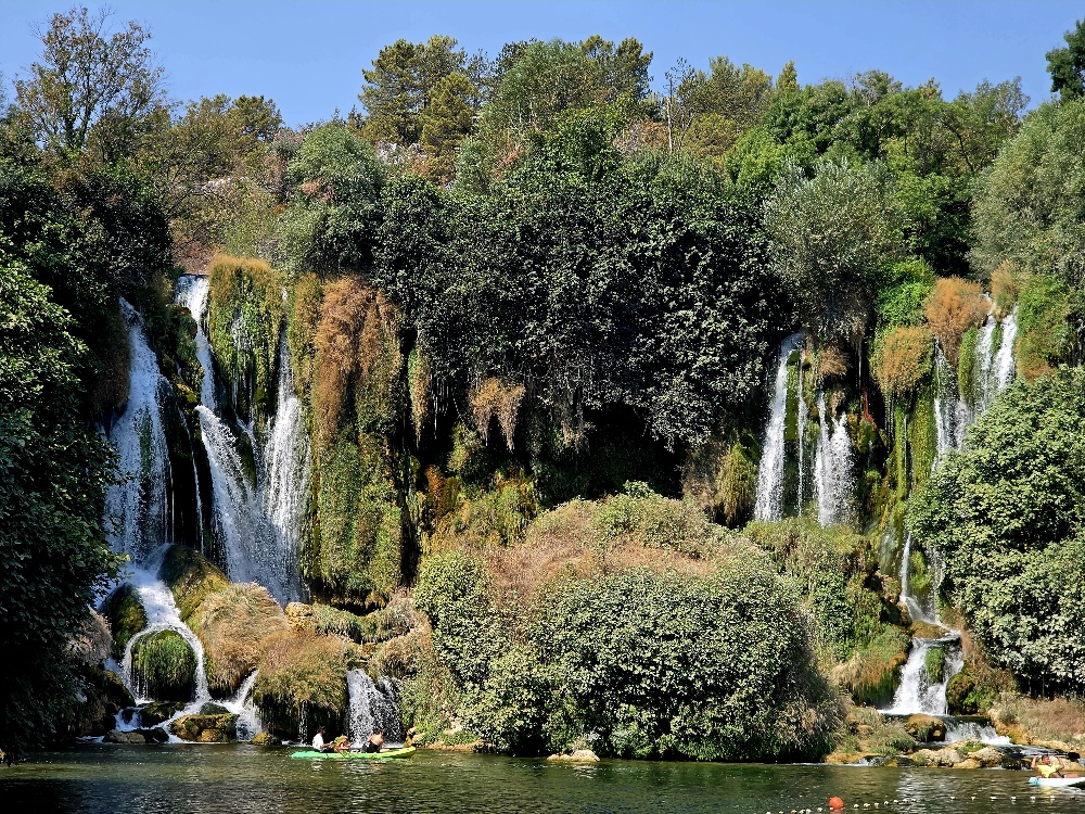 “Wide view of Kravica Falls, showing numerous waterfalls flowing over a lush, tufa-covered cliff into the swimming pool, with visitors kayaking and swimming below.”