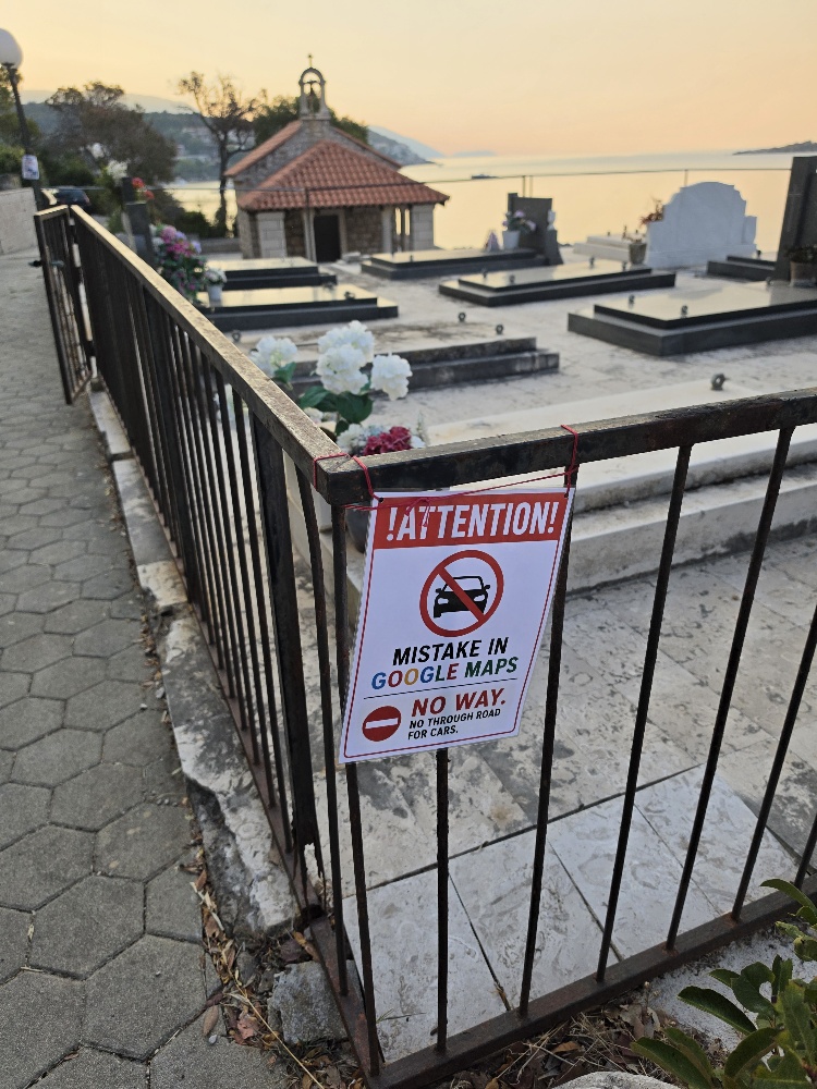 A small coastal cemetery with stone graves and white flowers, overlooking a calm bay at sunset. A sign on the foreground railing warns that Google Maps incorrectly directs cars by the cemetery.