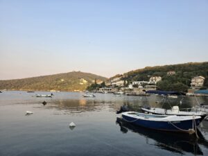 A calm coastal bay at sunset, with small fishing boats anchored in the water and houses scattered along the wooded hillsides.