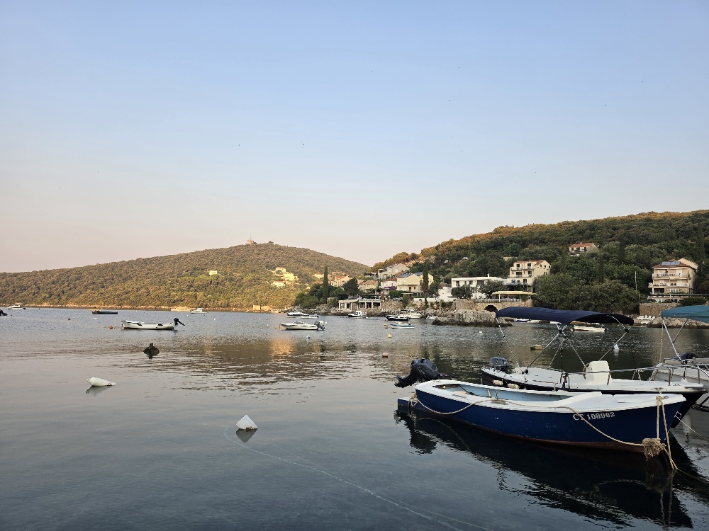 A calm coastal bay at sunset, with small fishing boats anchored in the water and houses scattered along the wooded hillsides.
