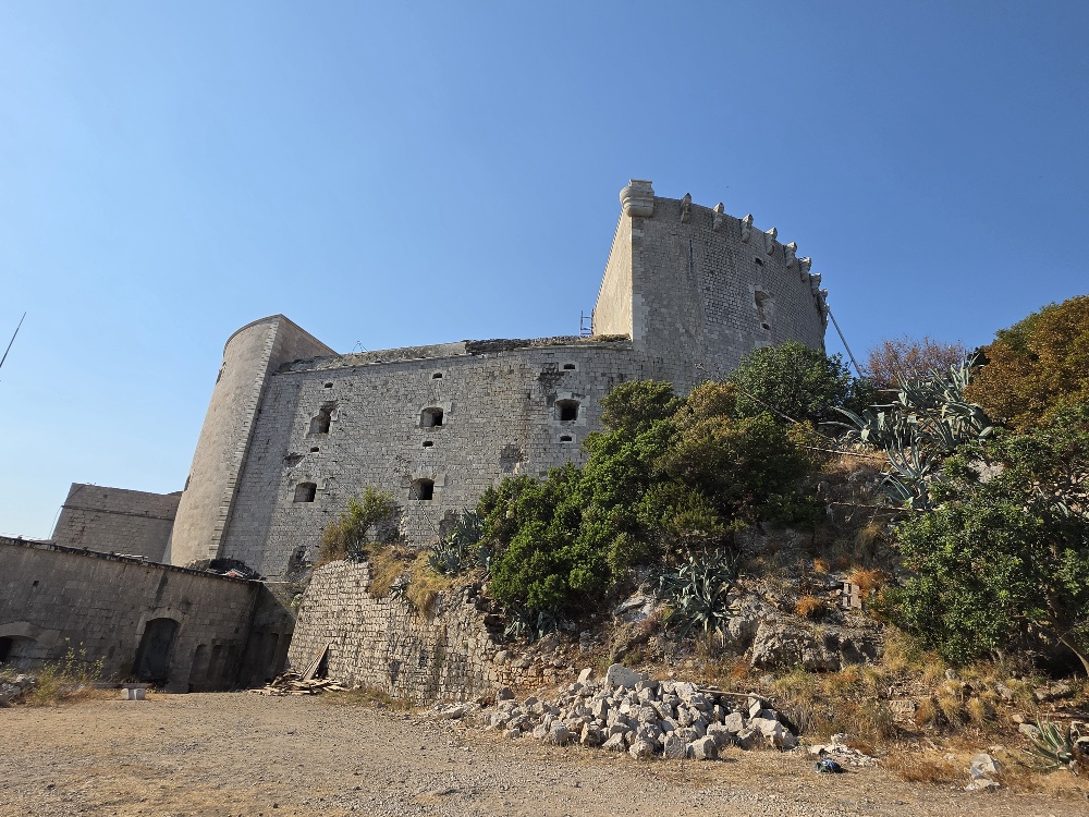 A large weathered stone fortress on a rocky hillside, with thick defensive walls, small openings and battlements surrounded by Mediterranean vegetation.