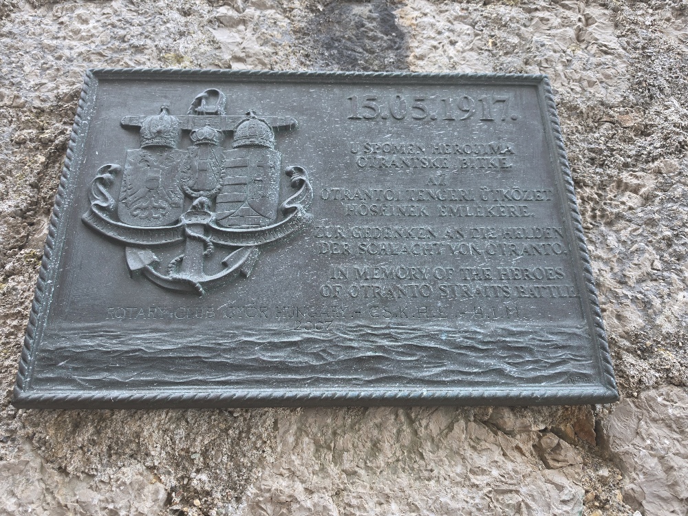 A dark metal memorial plaque mounted on a stone fortress wall, dated 15.05.1917, commemorating heroes of the Battle of Otranto Straits. The text reads: 15.05.1917. “USPOMENA HEROJIMA OTRANSKE BITKE DER SCHLACHT VON OTRANTO The German section appears to read: ZUR GEDENKEN AN DIE HELDEN DER SCHLACHT VON OTRANTO The English section reads: IN MEMORY OF THE HEROES OF OTRANTO STRAITS BATTLE At the bottom: ROTARY CLUB GYOR HUNGARY – CsK H.L. - H. I. M. – 2007 The Croatian heading is clearly “USPOMENA HEROJIMA OTRANSKE BITKE”, meaning