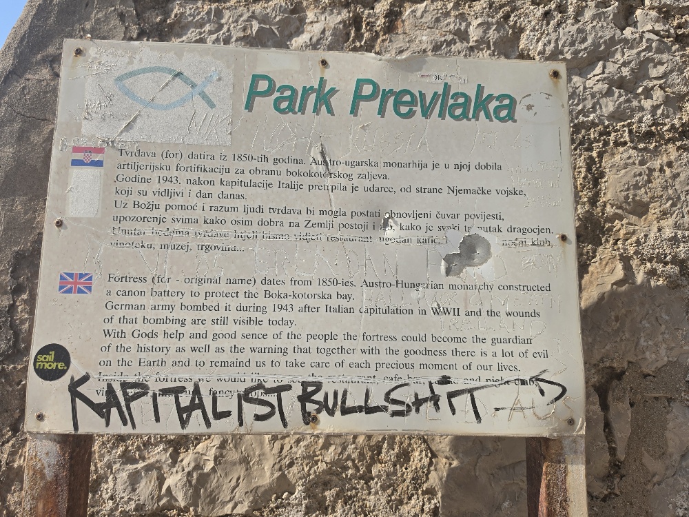 Sign that says PARK PREVLAKA in green and then in black by a flag of Croatia: Tvrđava (fort) datira iz 1850-tih godina. Austro-ugarska monarhija je u njoj dobila artiljerijsku fortifikaciju za obranu bokokotorskog zaljeva. Godine 1943. nakon kapitulacije Italije pretrpjela je udarce, od strane Njemačke vojske, koji su vidljivi i dan danas. Uz Božju pomoć i razum ljudi tvrđava bi mogla postati obnovljeni čuvar povijesti, upozorenje svima kako osim dobra na Zemlji postoji i zlo, kako je svaki trenutak dragocjen. [The remainder is heavily damaged/obscured, but appears to mention a restaurant, café, hotel, wine cellar, museum and shops.] English, next to a UK flag: Fortress (fcr - original name) dates from 1850-ies. Austro-Hungarian monarchy constructed a canon battery to protect the Boka-kotorska bay. German army bombed it during 1943 after Italian capitulation in WWII and the wounds of that bombing are still visible today. With Gods help and good sense of the people the fortress could become the guardian of the history as well as the warning that together with the goodness there is a lot of evil on the Earth and to remind us to take care of each precious moment of our lives. The final paragraph is substantially obscured by damage/graffiti, but I can make out references to the fortress becoming a restaurant, café, hotel, wine cellar, museum, shops... There is also a large handwritten/graffiti inscription across the sign that appears to read: KAPITALIST BULLSHIT 